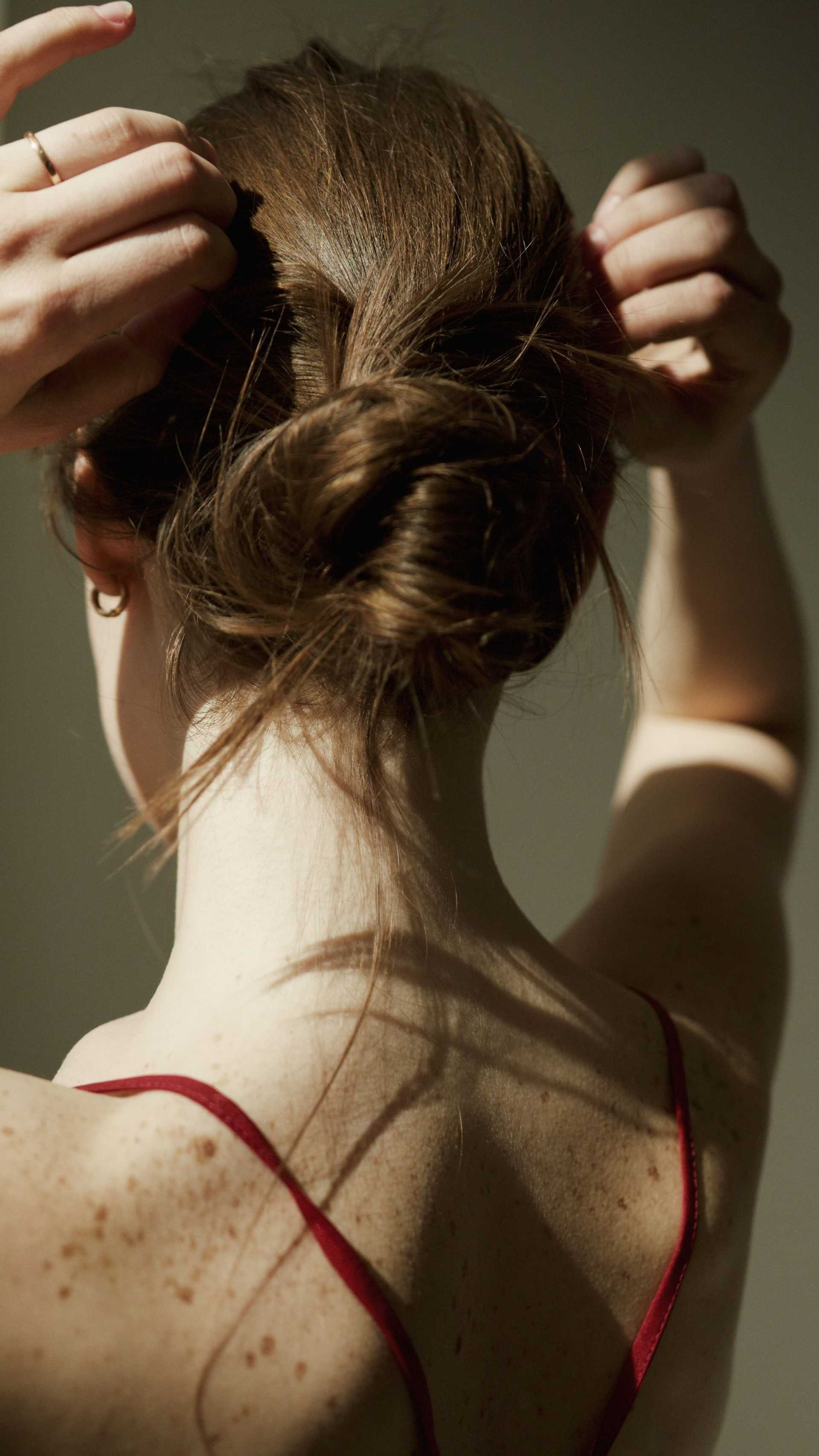 Woman styling hair in a bun, wearing a red strap dress, lit by soft, natural light.